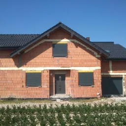 Surowy dom jednorodzinny w trakcie budowy z zamontowanymi roletami zewnętrznymi w kolorze grafitowym, widoczne ocieplenie ścian i brama garażowa.