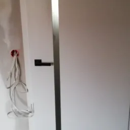 Białe drzwi wewnętrzne z czarną klamką i srebrnym paskiem, widoczne niedokończone wykończenie ściany z przewodami elektrycznymi.