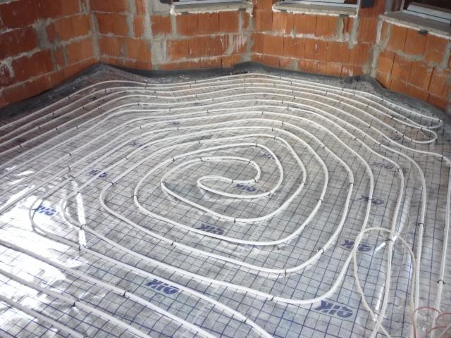 Rozłożone rury ogrzewania podłogowego na siatce montażowej, widoczne niedokończone ściany z cegły.