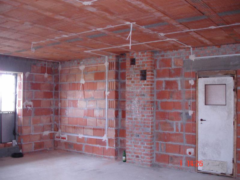 Surowy stan wnętrza z cegły z widoczną instalacją elektryczną natynkową, kable zabezpieczone uchwytami. Widoczna butelka po piwie na podłodze.