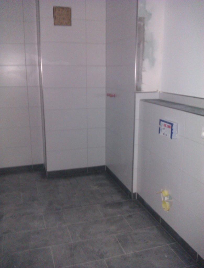 Wnętrze łazienki w trakcie wykończenia: białe płytki ścienne z szarą fugą, ciemnoszara podłoga z płytek, widoczne instalacje hydrauliczne i otwór po puszce elektrycznej.