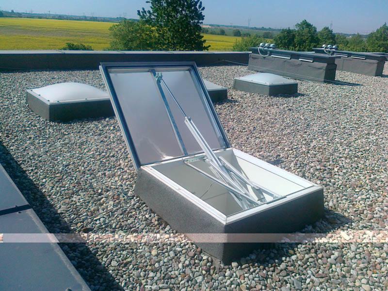 Otwarty świetlik dachowy na płaskim dachu pokrytym drobnym żwirem, widoczne mechanizmy otwierania, w tle inne świetliki i pole uprawne.
