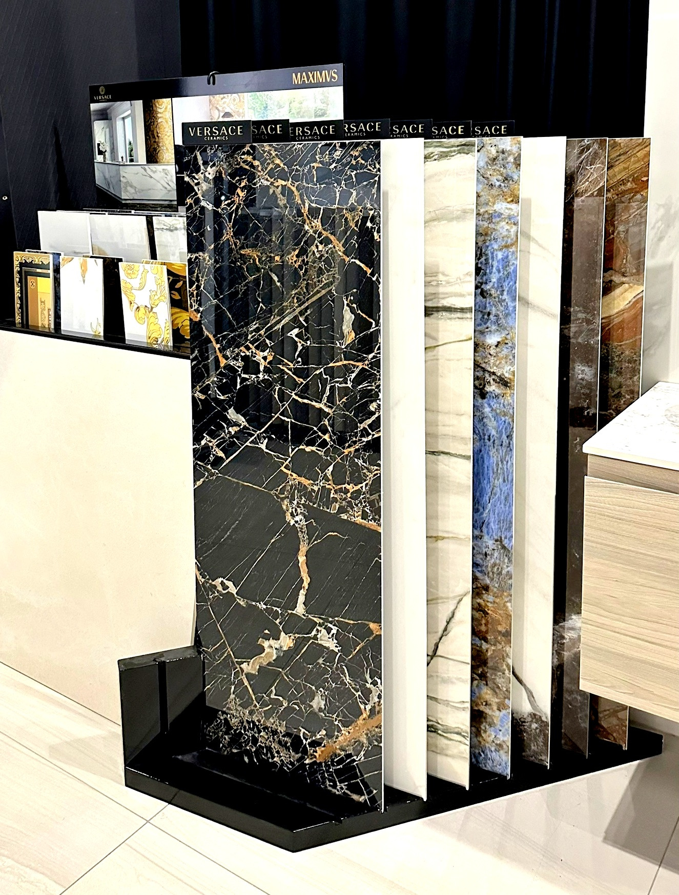 Ekspozycja pionowych próbek płytek ceramicznych, w tym czarnej z wyraźnym złotym żyłowaniem, w salonie łazienek; widoczne logo Versace na niektórych płytkach.