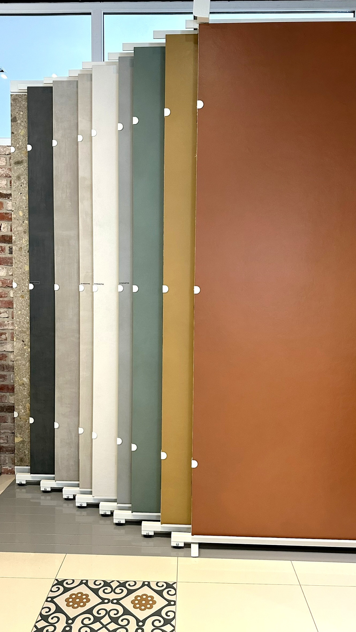 Ekspozycja próbek płytek wielkoformatowych w różnych kolorach i fakturach, ustawionych pionowo na białych stojakach, na pierwszym planie dekoracyjna płytka podłogowa.