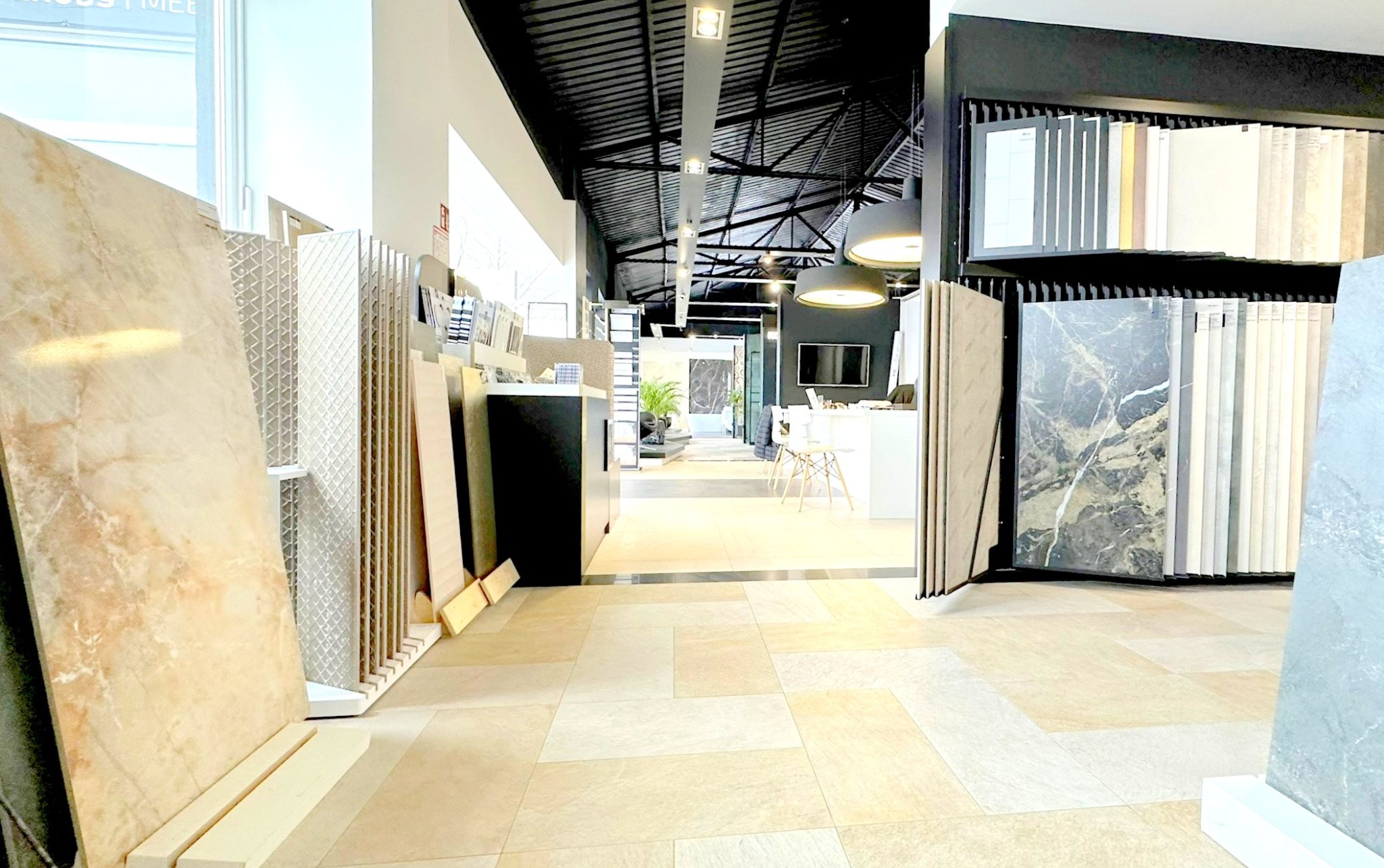 Ekspozycja płytek ceramicznych w salonie: różnorodne wzory i kolory, imitacja kamienia, gresy, oświetlenie sufitowe, widok na dalszą część sklepu z meblami łazienkowymi i roślinami.