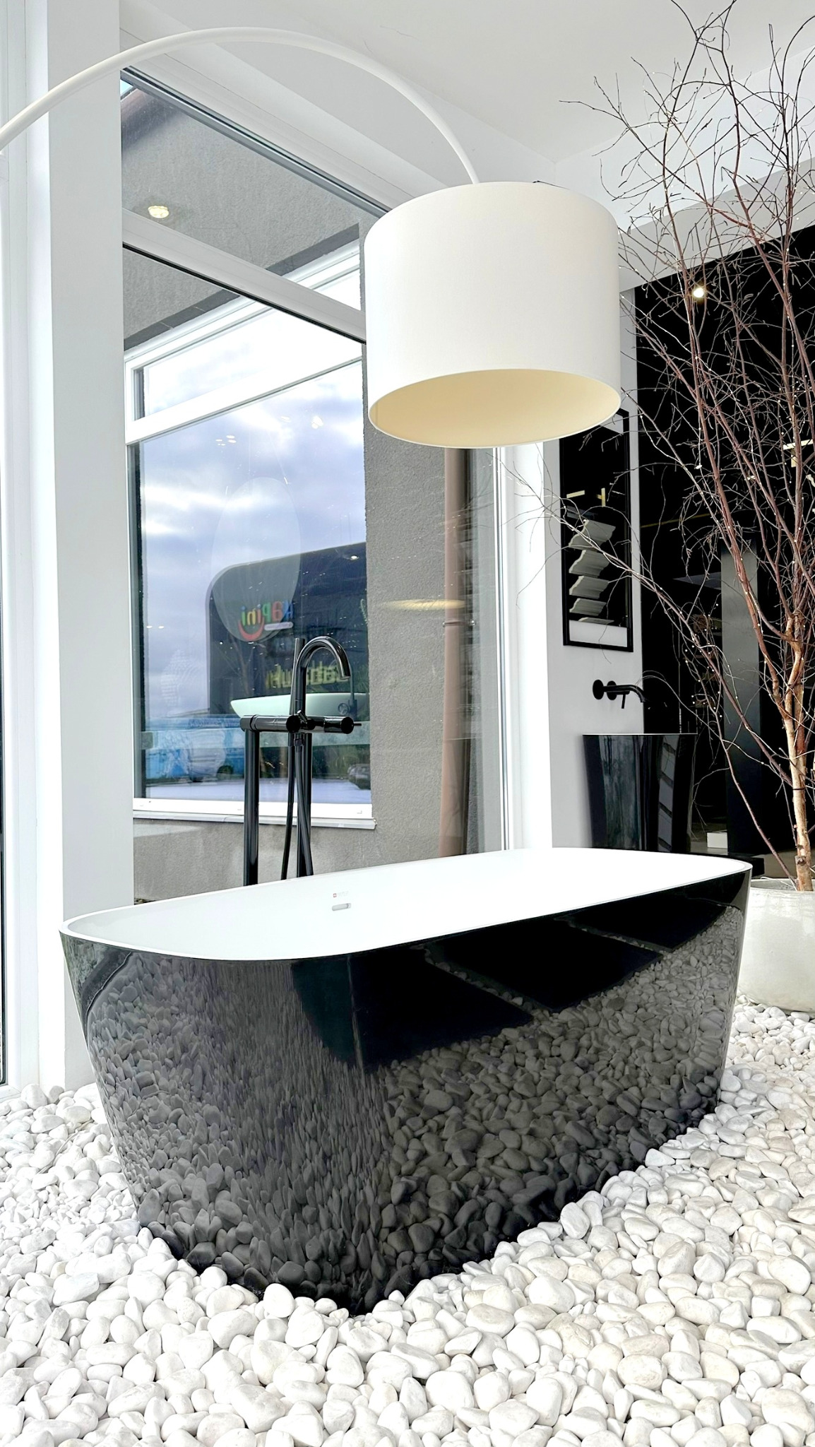 Wolnostojąca wanna w czarnym kolorze, otoczona białymi otoczakami, podświetlona lampą z abażurem, z minimalistyczną armaturą w tle i dekoracyjnymi gałązkami.