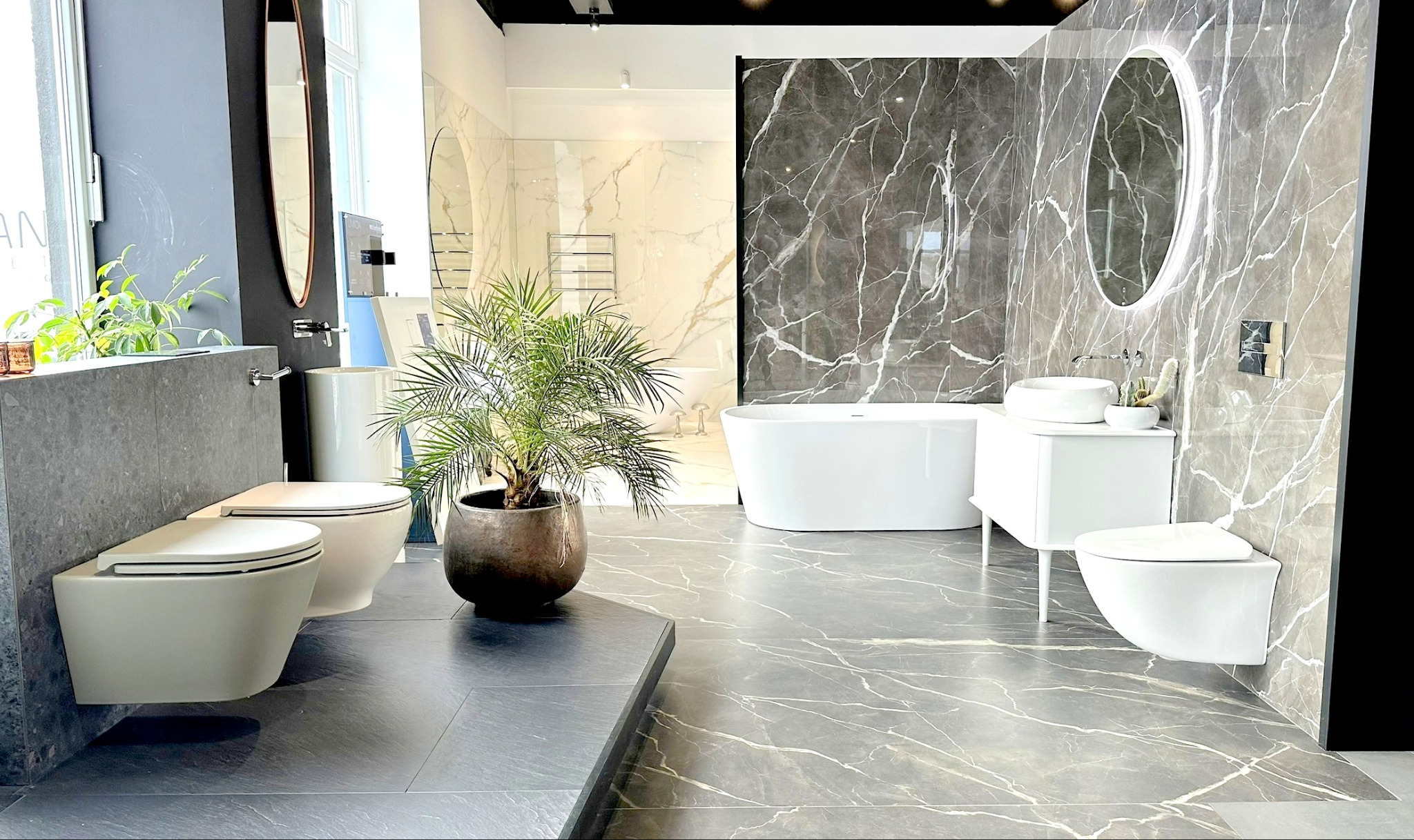 Salon łazienek prezentuje aranżację z wolnostojącą wanną, umywalką nablatową, podwieszanymi toaletami oraz szarą marmurową płytką na ścianach i podłodze. Centralnym elementem jest duża roślina...