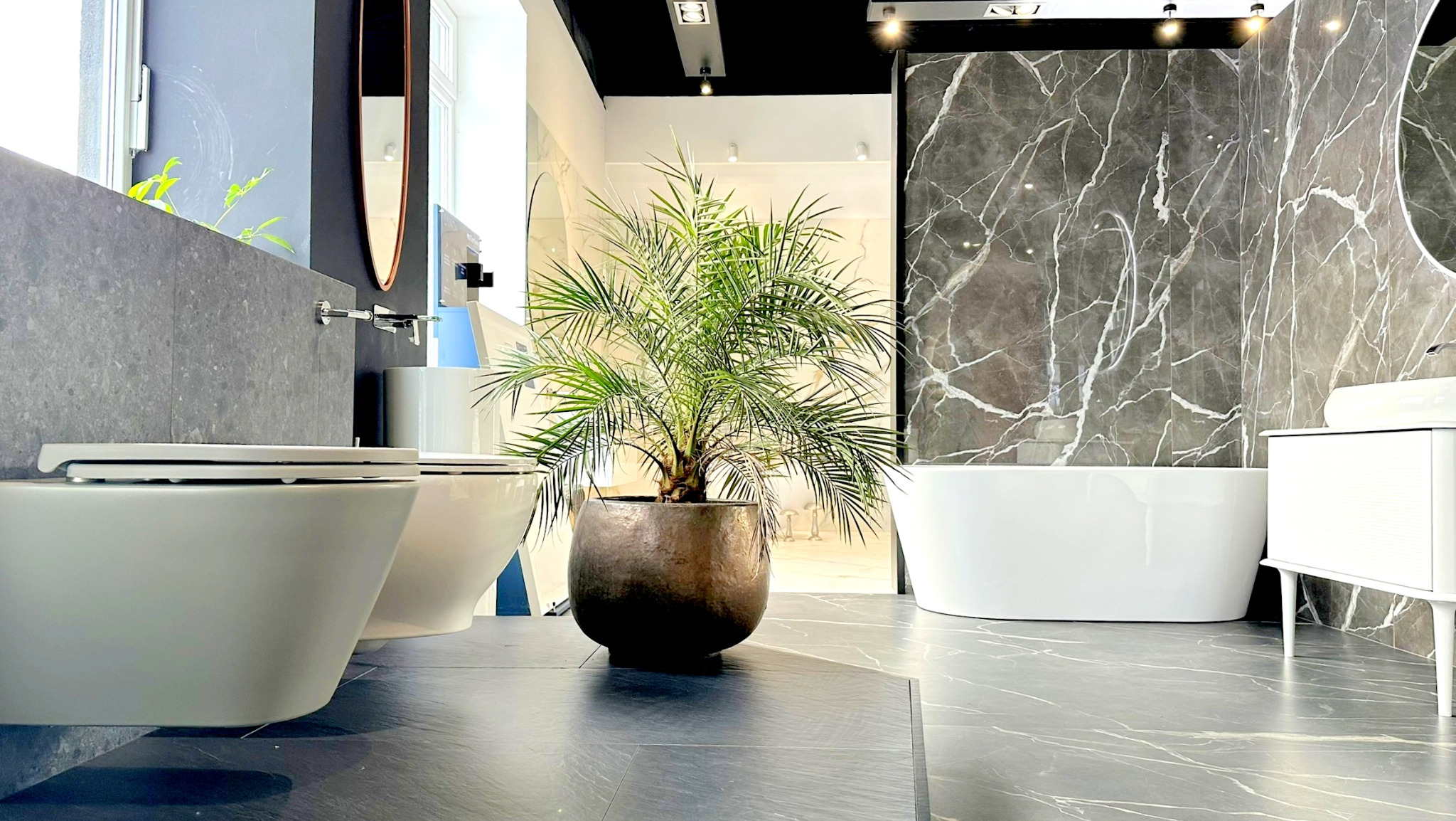 Nowoczesna aranżacja łazienki z dwoma podwieszanymi toaletami, wolnostojącą wanną na tle marmurowej ściany i palmą w metalowej donicy na podłodze z szarego kamienia.