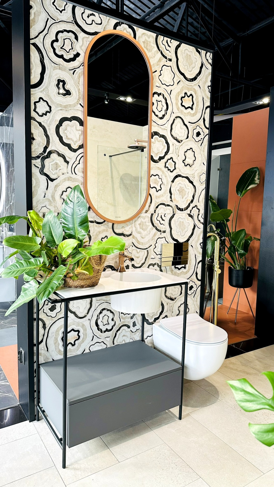 Aranżacja łazienki z umywalką nablatową, lustrem w miedzianej ramie, toaletą podwieszaną i szafką w kolorze szarym, uzupełniona roślinami doniczkowymi i płytkami w geometryczny wzór.