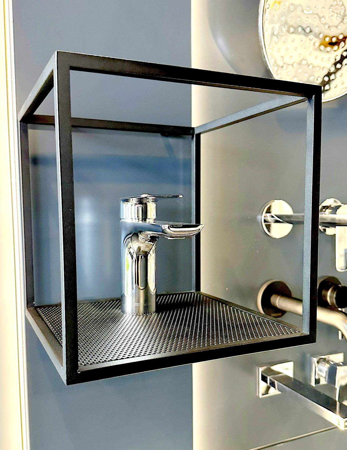 Błyszcząca chromowana bateria umywalkowa prezentowana w czarnej, minimalistycznej, kubicznej ramie z perforowanym dnem, na tle ekspozycji armatury łazienkowej.