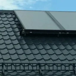 Dwa panele solarne zamontowane na czarnym, metalowym dachu z dachówki ceramicznej, widoczne rury i uchwyty mocujące, błękitne niebo w tle.