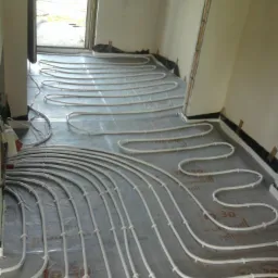 Białe rury ogrzewania podłogowego ułożone na folii izolacyjnej w surowym wnętrzu, widoczne mocowania i fragmenty ścian.