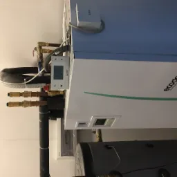 Biała pompa ciepła Mitsubishi Ecodan z widocznym sterownikiem, rurami miedzianymi i zbiornikiem buforowym na tle białej ściany.