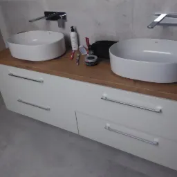 Nowoczesna łazienka z dwoma owalnymi umywalkami nablatowymi, drewnianym blatem i białymi szafkami z poziomymi, metalowymi uchwytami, widoczne kosmetyki i akcesoria do higieny osobistej.