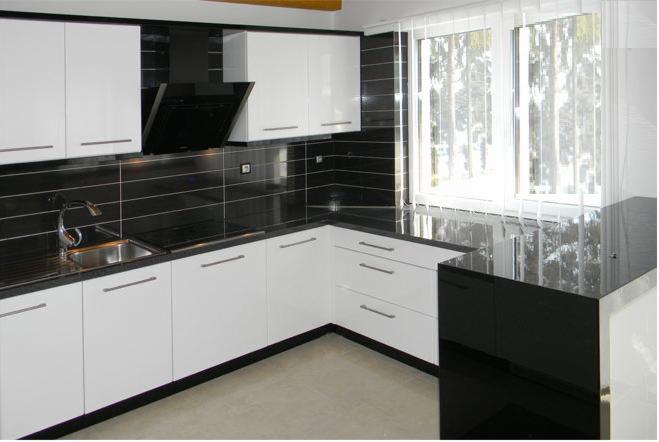 Nowoczesna kuchnia z białymi szafkami i czarnym blatem, czarne płytki na ścianie, okno z żaluzjami pionowymi.