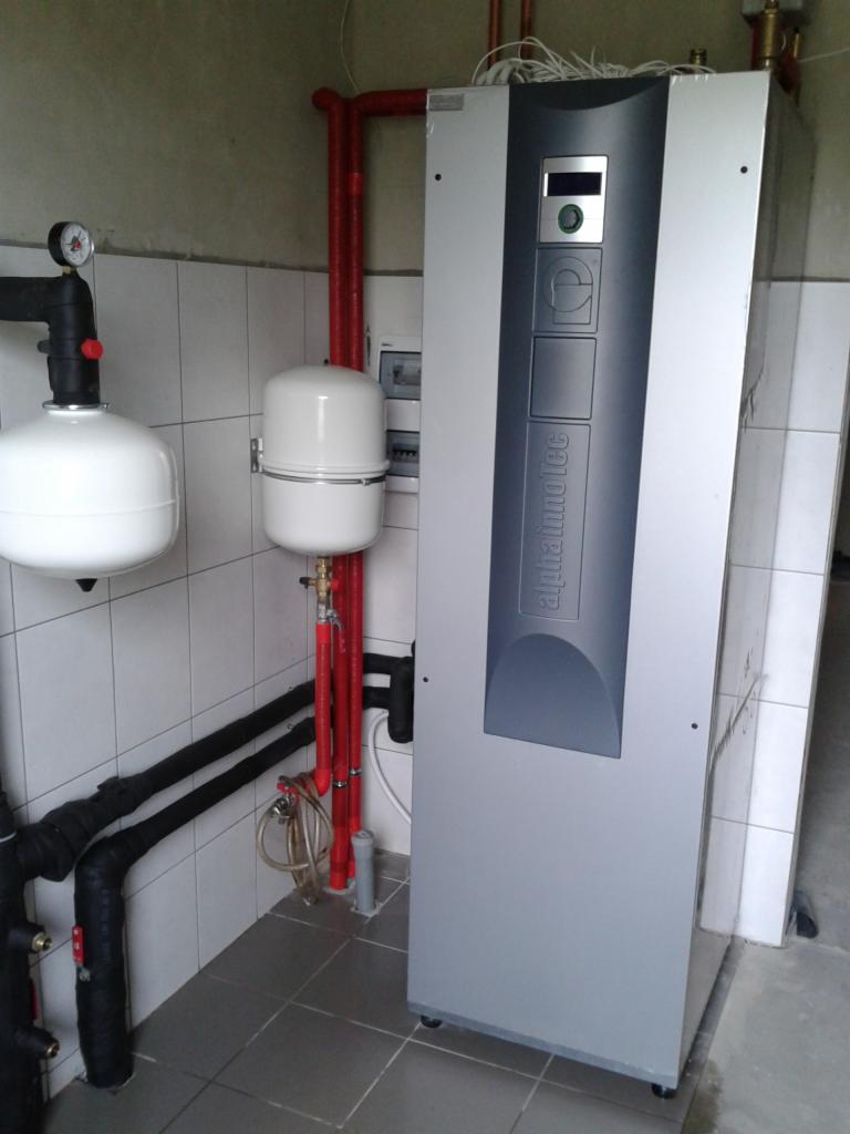 Zamontowany, nowoczesny system grzewczy Alpha Inno Tec z białą obudową, czerwonymi rurami i białymi zbiornikami w pomieszczeniu wyłożonym białymi kaflami.
