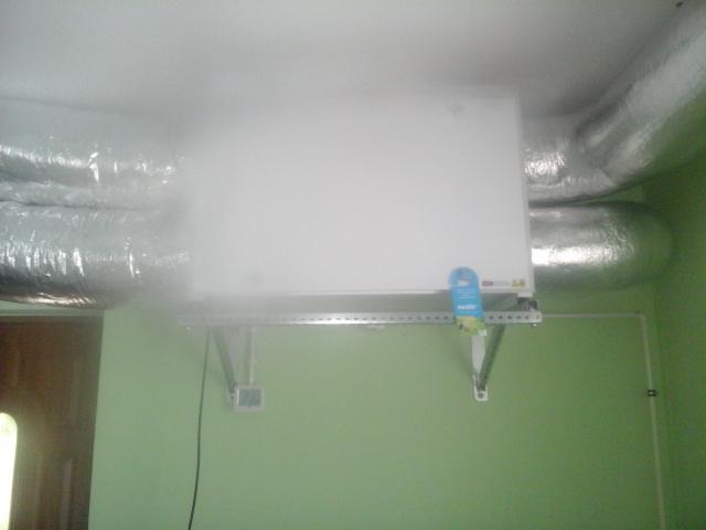 Zamontowana centrala rekuperacyjna z widocznymi przewodami wentylacyjnymi w pomieszczeniu w Wieluniu. Biała obudowa urządzenia na zielonej ścianie.