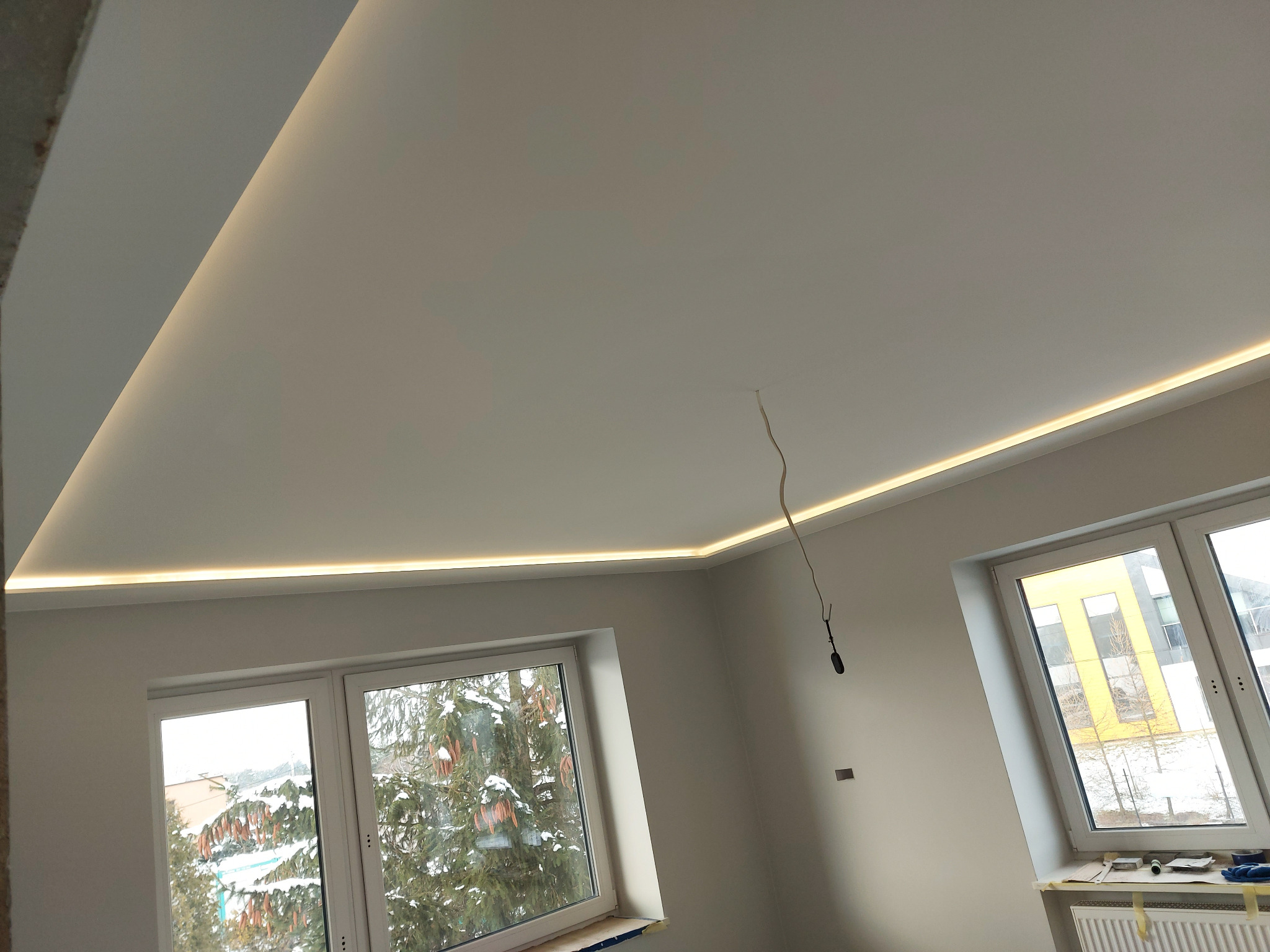 Nowoczesne oświetlenie LED wzdłuż sufitu w jasnym pokoju z dwoma oknami, widok na ośnieżone drzewa i budynek za oknem, kabel zwisający z sufitu.