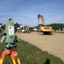 Zielony teodolit na pomarańczowo-żółtym statywie ustawiony na trawie, w tle plac budowy z koparkami i pracownikami, niebieskie niebo.