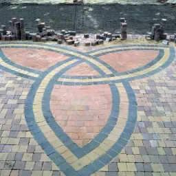 Wzór triskelionu ułożony z kostki brukowej w odcieniach brązu, beżu i grafitu, w tle przygotowany teren pod dalsze prace.