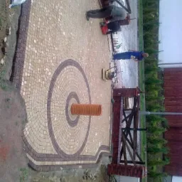 Układanie jasnobrązowej kostki brukowej w okrągły wzór wokół rury kanalizacyjnej; pracownicy w trakcie montażu obrzeży z cegły i drewnianego płotu.