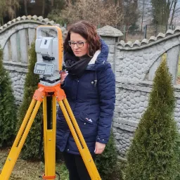 Kobieta w okularach i niebieskiej kurtce obsługuje żółty teodolit ZTS Cygnus na trójnogu, na tle betonowego płotu i iglaków.