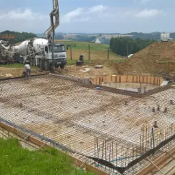 Przygotowanie zbrojenia fundamentów pod budowę domu; widoczne szalunki, siatka zbrojeniowa, węzły instalacyjne oraz betonowozy w tle.