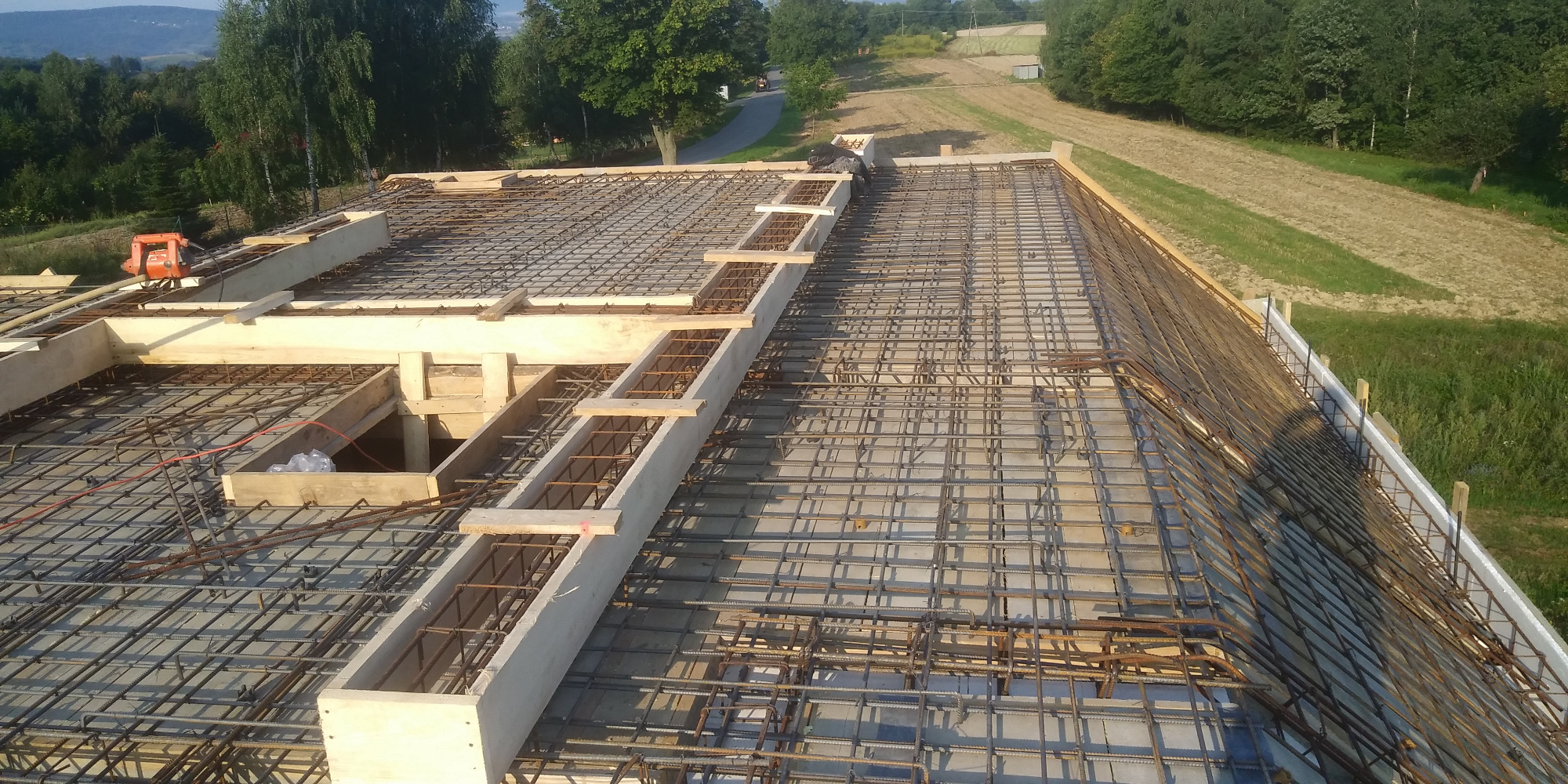 Zbrojenie stropu budynku w trakcie budowy, widoczne drewniane szalunki i siatka z prętów zbrojeniowych na tle zieleni.