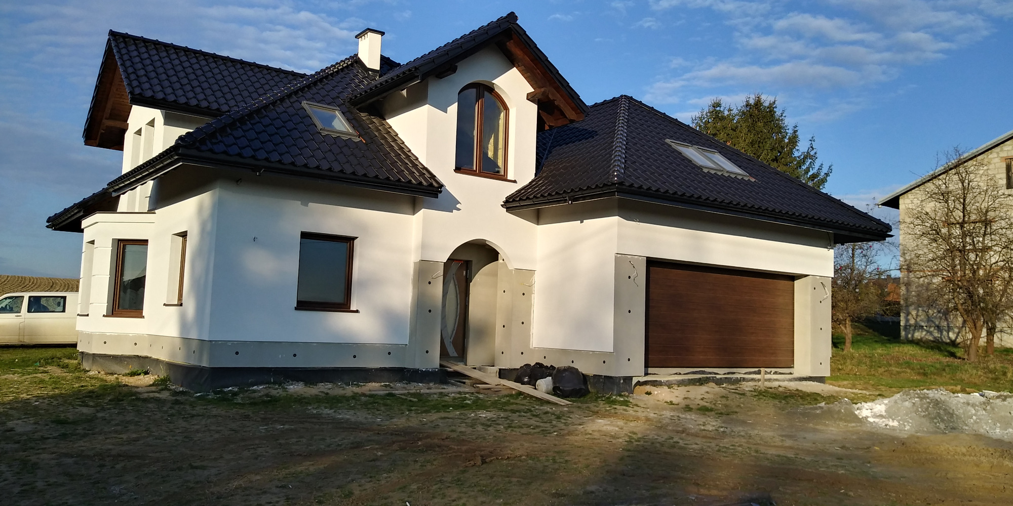 Niedokończony, biały dom z ciemną dachówką, widoczne fragmenty elewacji przed wykończeniem, boczny garaż z brązową bramą, teren wokół w trakcie prac ziemnych, miejscowość Korczyna.