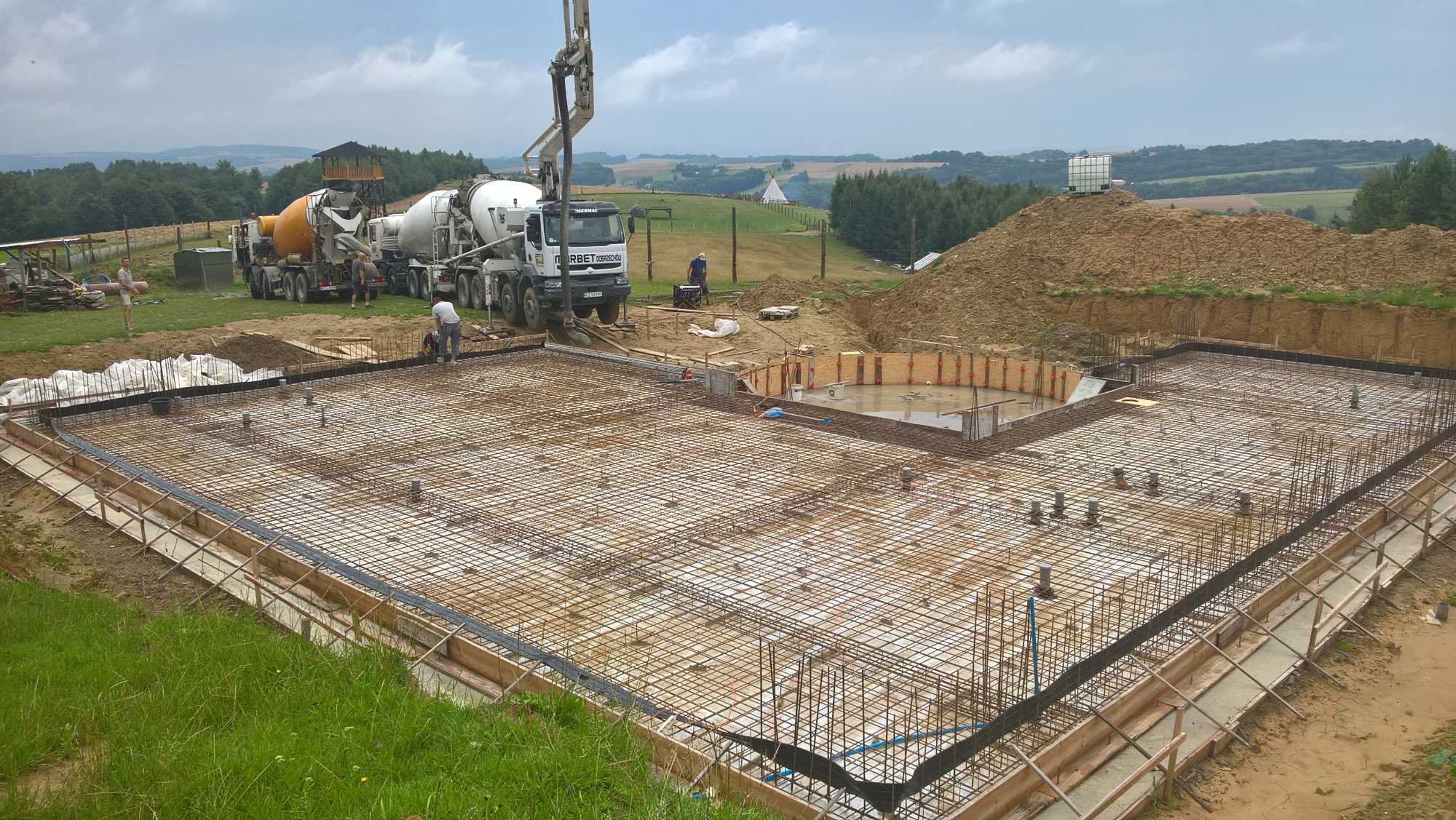 Przygotowanie zbrojenia fundamentów pod budowę domu; widoczne szalunki, siatka zbrojeniowa, węzły instalacyjne oraz betonowozy w tle.