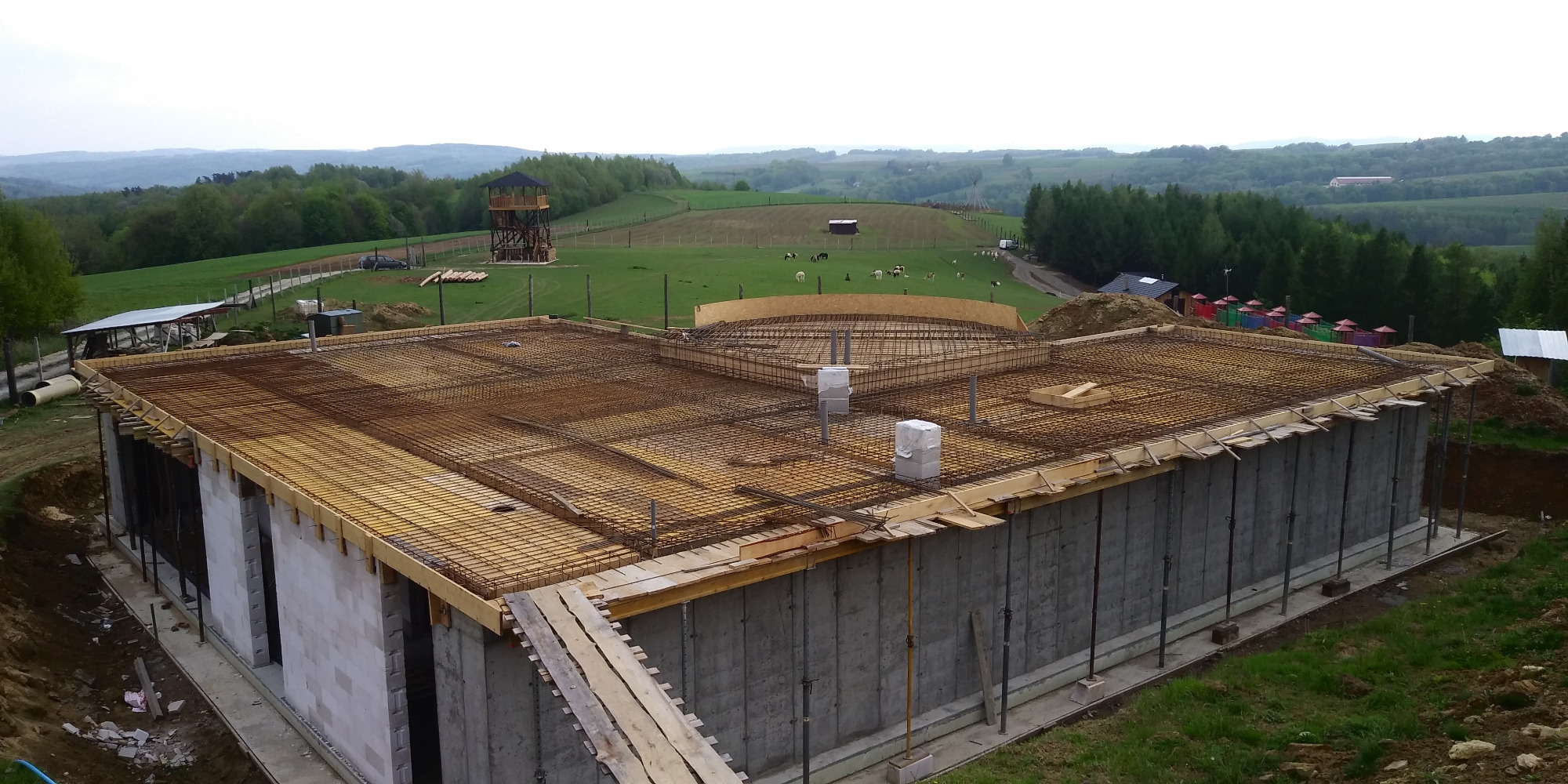 Wznoszona konstrukcja żelbetowa z widocznym zbrojeniem i szalunkiem, na tle malowniczego krajobrazu z wieżą widokową i pasącymi się zwierzętami.