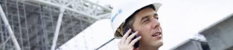Mężczyzna w białym kasku z logo firmy ubezpieczeniowej rozmawia przez telefon na tle konstrukcji stalowej.