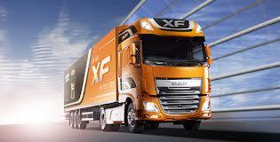 Pomarańczowy ciągnik siodłowy DAF XF z naczepą na tle błękitnego nieba i metalowych konstrukcji.