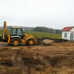Żółta koparko-ładowarka JCB przygotowuje wykop pod konstrukcję studni kręgowej z białym domkiem z czerwoną dachówką w tle, na rozjeżdżonym, błotnistym terenie.