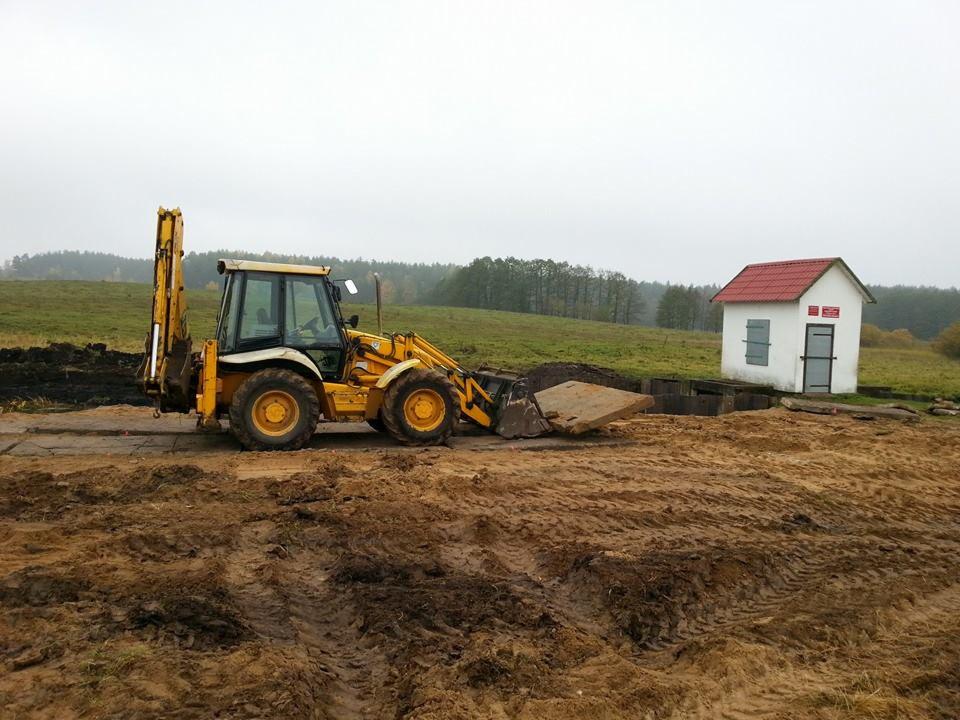 Żółta koparko-ładowarka JCB przygotowuje wykop pod konstrukcję studni kręgowej z białym domkiem z czerwoną dachówką w tle, na rozjeżdżonym, błotnistym terenie.