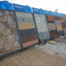 Ekspozycja różnorodnych wzorów kostki betonowej marki Polbruk, prezentowana na metalowych stojakach na zewnątrz, z ułożonymi próbkami na ziemi.