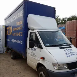 Samochód dostawczy z plandeką z reklamą składu budowlanego RODA na tle palet ze styropianem Austrotherm i cegłami Wienerberger Porotherm.