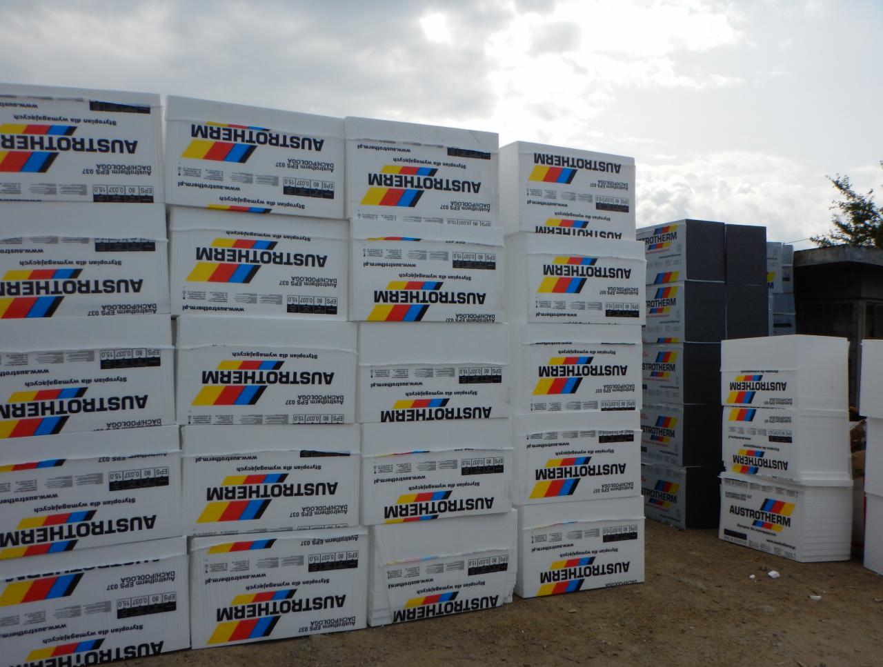 Stosy białych paczek styropianu Austrotherm EPS 037 DACH/PODŁOGA, ułożone na placu budowy, widoczne szare płyty styropianowe w tle, pochmurne niebo.