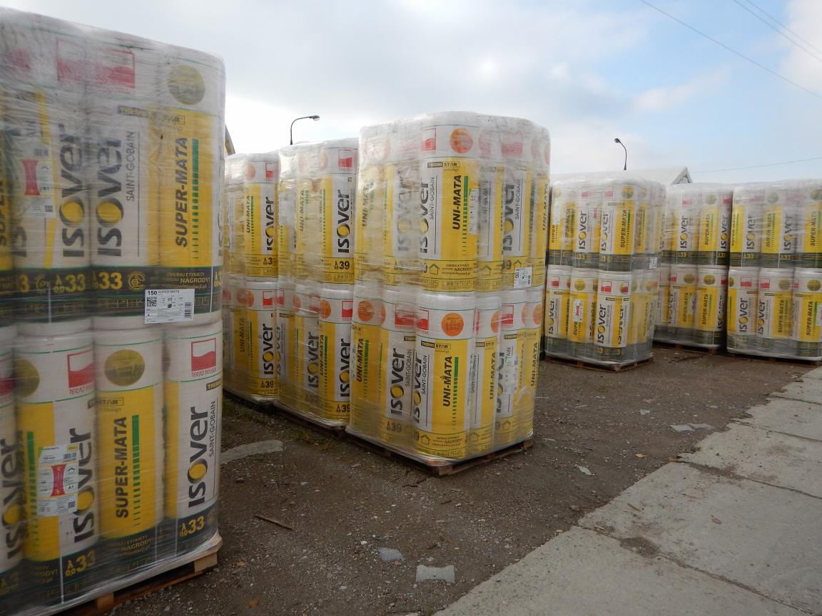 Palety z żółto-białymi rolkami materiału izolacyjnego Isover Super-Mata i Uni-Mata, owinięte folią, składowane na zewnątrz na betonowym podłożu, widoczne słupy oświetleniowe w tle.