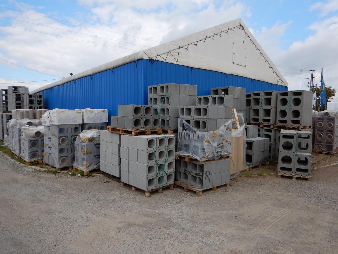 Magazyn zewnętrzny z szarymi blokami betonowymi ułożonymi na paletach, część zafoliowana, na żwirowym podłożu, w tle niebieski magazyn z białym dachem.