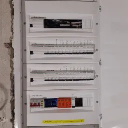 Rozdzielnica elektryczna w białej obudowie, widoczne wyłączniki różnicowoprądowe Schneider Electric z opisami obwodów (indukcja piętro, indukcja parter) oraz wyłącznik główny Ideal...