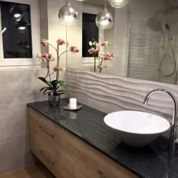 Nowoczesna łazienka z drewnianą szafką pod umywalką, czarnym blatem i okrągłą umywalką nablatową. Nad blatem wiszą trzy srebrne lampy.