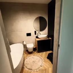 Nowoczesna łazienka z owalną wanną, okrągłym lustrem nad umywalką nablatową i drewnopodobną podłogą, widziana z perspektywy wejścia.