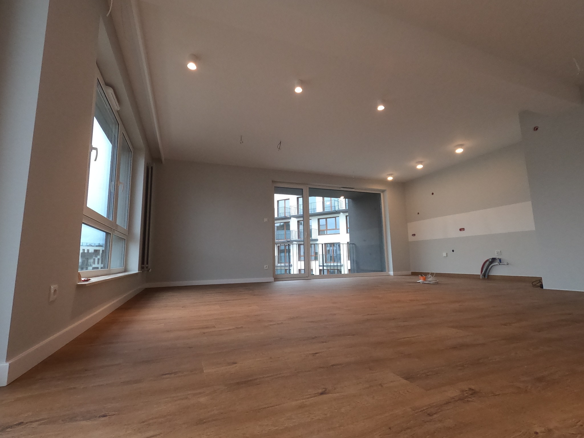 Przestronne wnętrze mieszkania w stanie deweloperskim z drewnopodobną podłogą, dużymi oknami i przygotowanymi instalacjami elektrycznymi na ścianach.