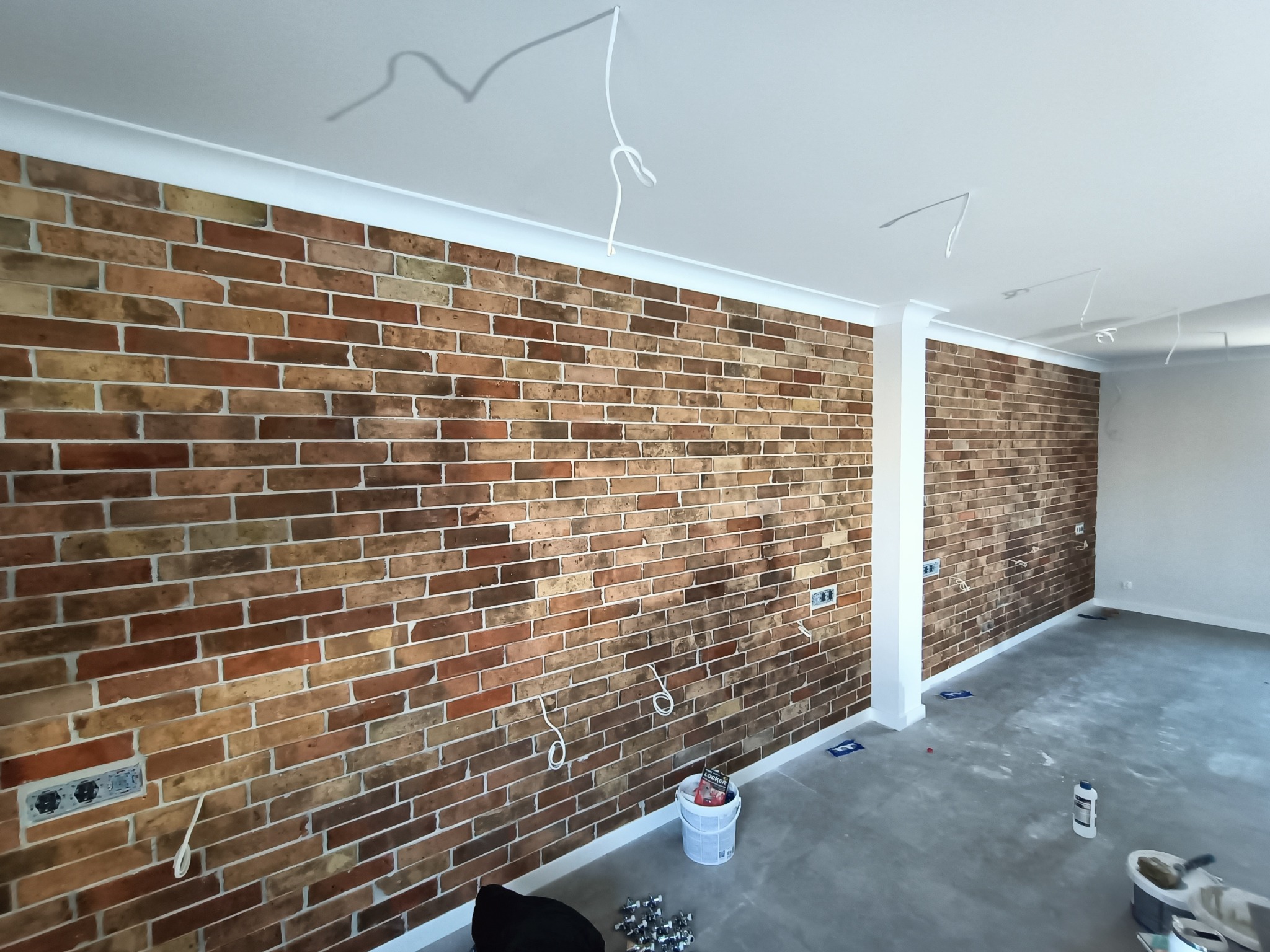 Ściana z cegły klinkierowej w odcieniach brązu i beżu, z widocznymi puszkami elektrycznymi i wystającymi kablami, biała listwa przypodłogowa, surowa wylewka na podłodze, biały sufit z wystającymi...