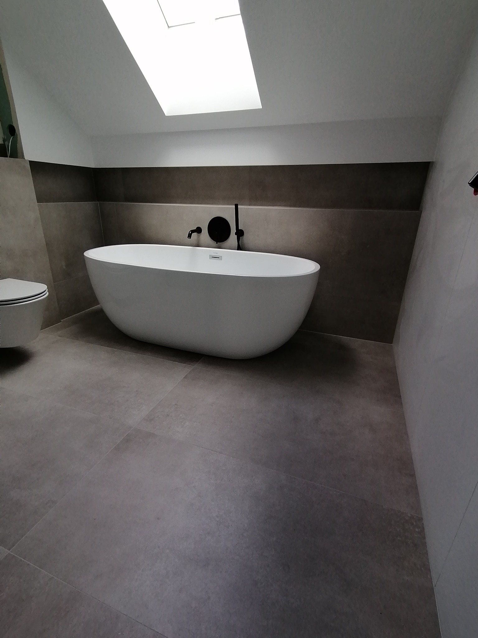 Nowoczesna łazienka na poddaszu z wolnostojącą białą wanną, czarną armaturą i szarą płytką podłogową, oświetlona przez okno dachowe.