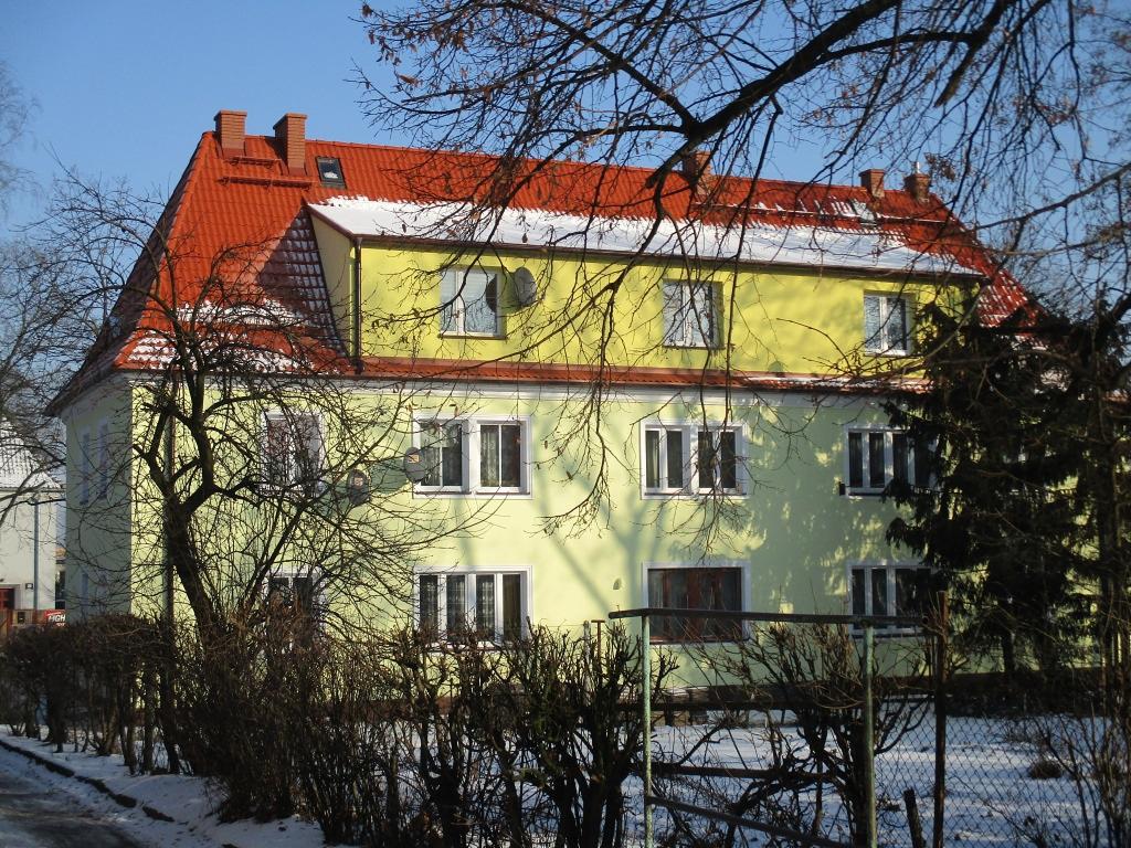 Dwupiętrowy budynek mieszkalny w Ciechanowie, z żółtą elewacją, czerwonym dachem i białymi ramami okiennymi, częściowo zasłonięty nagimi gałęziami drzew w zimowej scenerii.