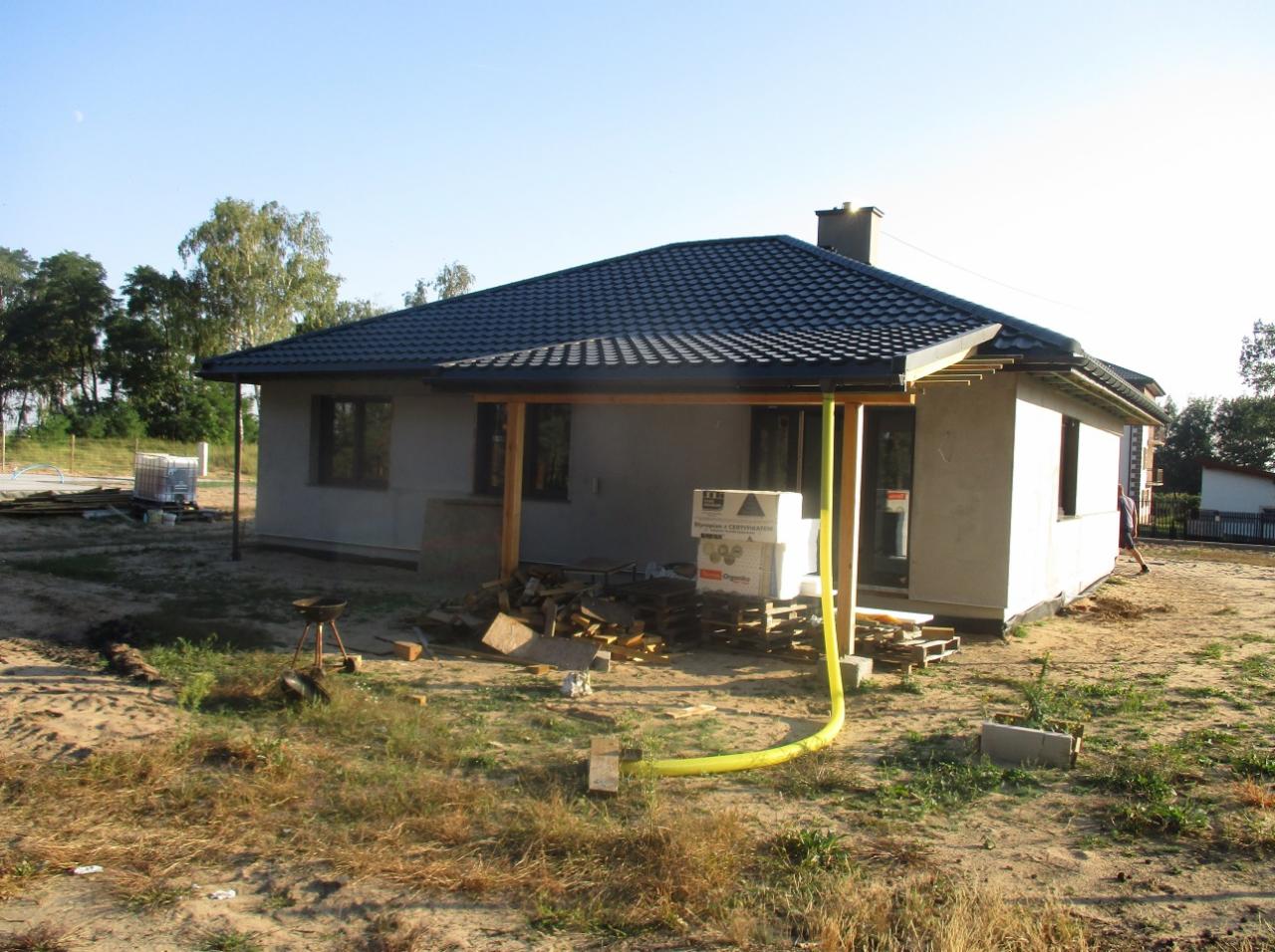 Niedokończony dom parterowy z ciemnoszarą dachówką, widoczne drewniane podpory zadaszenia, żółty wąż na trawniku, materiały budowlane i grill na działce.