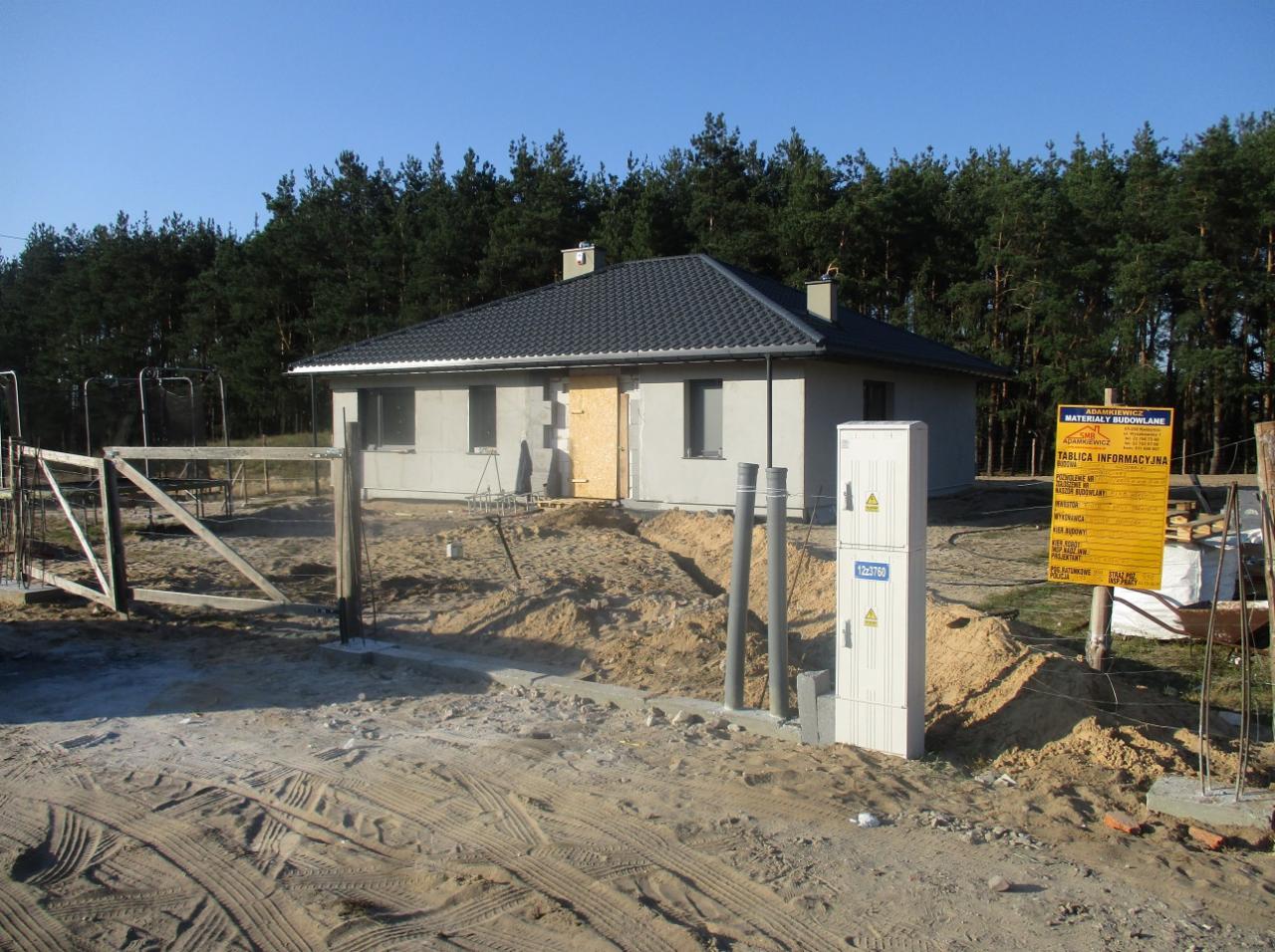 Niedokończony parterowy dom z szarą dachówką i surową elewacją, widoczna skrzynka elektryczna, teren wokół w stanie budowy, tablica informacyjna budowy w tle lasu.