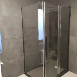 Nowoczesna kabina prysznicowa z czarnymi profilami i deszczownicą, szare płytki imitujące marmur na ścianach, widoczny grzejnik drabinkowy i odpływ liniowy.